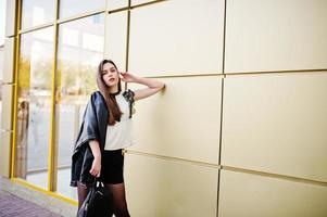 Elegante chica morena vestida con chaqueta de cuero y pantalones cortos con mochila contra la pared dorada. foto