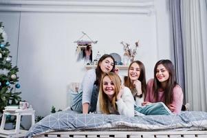 Four cute friends girls wear on warm sweaters and black pants on bed at new year decorated room on studio, play with pillows. photo