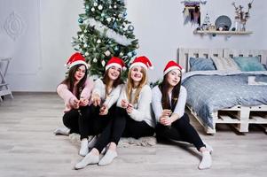 cuatro lindas amigas usan suéteres cálidos, pantalones negros y sombreros rojos de santa contra el árbol de año nuevo con decoración navideña en la habitación blanca y sostienen luces de bengala en las manos. foto