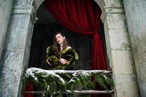 chica morena con abrigo de piel verde contra el viejo arco con columnas y cortinas rojas. foto