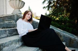 Good-looking young woman in white blouse, wide black pants and black classic high heels sitting on stairs and working on her laptop. photo