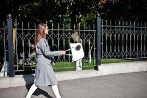 Girl in gray coat with sunglasses and handbag walking at street against iron fence. photo