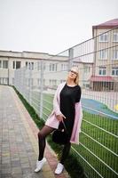 Blonde girl at glasses and pink coat, black tunic and handbag posed against fence at street. photo