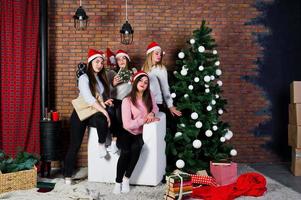 cuatro lindas amigas usan suéteres cálidos, pantalones negros y sombreros de santa contra el árbol de año nuevo con decoración navideña en el estudio. foto