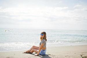 hermosa modelo relajándose en una playa de mar, usando jeans cortos, camisa de leopardo y gafas de sol. foto