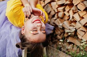 Young funny girl with bright make-up, like fairytale princess, wear on yellow shirt and violet skirt lying against wooden background. photo