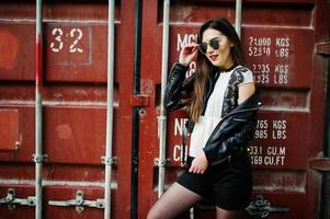 Stylish brunette girl wear on leather jacket and shorts, sunglasses against shipping container. photo