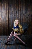 Elegante blonde girl wear on fur coat sitting on chair at studio against wooden background. photo