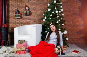 ropa de niña linda en suéteres cálidos, pantalones negros contra el árbol de año nuevo con decoración navideña en el estudio. foto