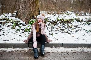 Stylish girl in fur coat and headwear at winter day on sitting border ofroad. photo