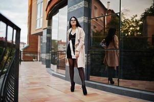 Black hair sexy woman in glasses and coat posed against building with modern windows. photo