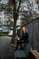 Blonde fashionable girl in long black leather coat posed on bench. photo