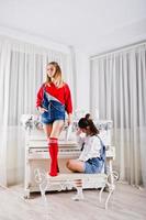 Two fun beautiful girls friends wear in overalls jeans shorts and gaiters against piano with new year decoration. photo