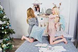 Two elegante blonde girl wear on warm tunic sitting on bed and play with pillows against new year tree. photo