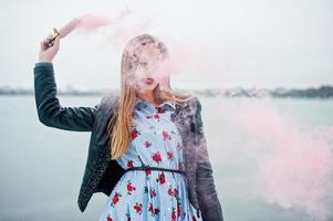 Stylish girl in leather jacket hold pink smoke flare at winter day against frozen lake. photo