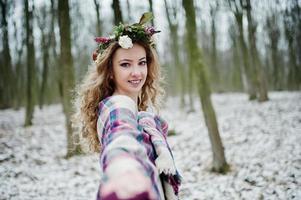 Curly cute blonde girl with wreath in checkered plaid at snowy forest in winter day. photo