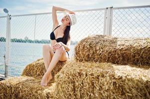 Portrait of a gorgeous girl in black bikini swimsuit posing on the hay bale with a hat by the lake. photo