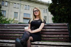 Elegant blonde girl wear on black posing at streets of town, sitting on bench with cup of coffee at hands. photo