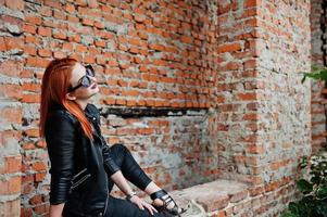 Red haired stylish girl in sunglasses wear in black, against abadoned place with brick walls. photo