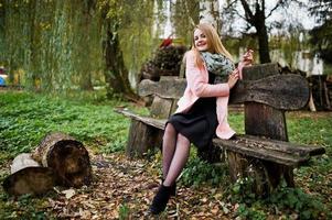 jovencita rubia con abrigo rosa sentada en un banco posada sobre un fondo de tocones de madera. foto