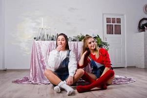 Two fun beautiful girls friends wear in overalls jeans shorts and gaiters against table with new year decoration. photo