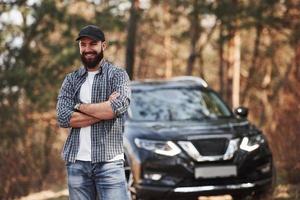 Smiling for the camera. Bearded man near his brand new black car in the forest. Vacations concept photo