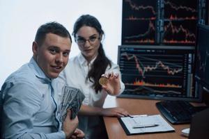 Showing off earned things. Beautiful woman and cheerful man holding bitcoin and money in hands while sitting in modern office with many monitors with graphs photo