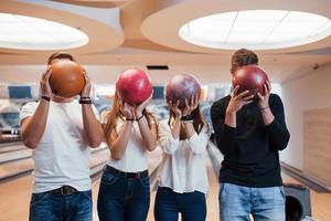 hiding faces behin the balls. Young cheerful friends have fun in bowling club at their weekends photo