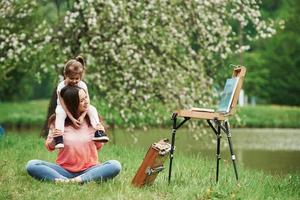 Enjoying by looking at painting. Mother and daughter sitting near easel in the park at spring time photo