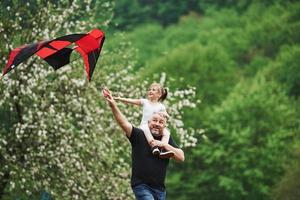 Spending weekend together. Running with red kite. Child sitting on the man's shoulders. Having fun photo