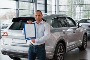 Serious businessman. Manager stands in front of modern silver colored car with paper and documents in hands photo