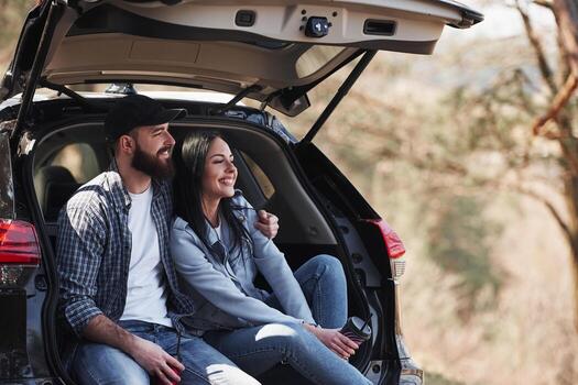 Toothy smiles. Sitting on rear part of automobile. Enjoying the nature. Couple have arrived to the forest on their brand new black car photo
