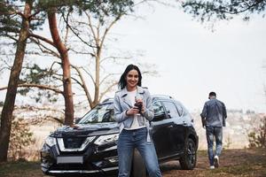 Holds cup with warm drink. Couple have arrived to the forest on their brand new black car photo