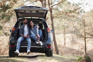 Having a break. Sitting on rear part of automobile. Enjoying the nature. Couple have arrived to the forest on their brand new black car photo