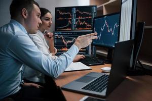 Man talks and shows on the numbers on monitor with hand. Team of stockbrokers are having a conversation in a office with multiple display screens photo