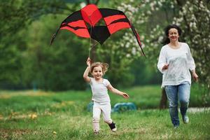 Fast and active. Positive female child and grandmother running with red and black colored kite in hands outdoors photo