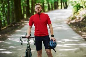 Holding safety helmet in hand. Cyclist on a bike is on the asphalt road in the forest at sunny day photo