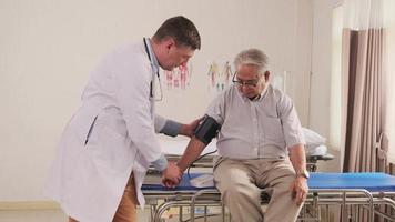 medico maschio caucasico in uniforme controlla la malattia paziente anziano, pressione sanguigna misurata nel letto del pronto soccorso nel reparto ospedaliero, clinica medica per anziani, consulente per l'esame doloroso. video