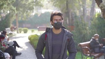College-Student, der in Zeitlupe im Park spazieren geht. junger Mann zu Fuß. Mann mit schwarzer Maske und Brille. pandemische Bedingungen. Alltag. Schüler mit Rucksack. video