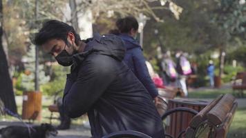 uomo dalla pelle scura seduto su una sedia nel parco. seduto su una sedia nel parco. persone che camminano con i cani. profondità di campo. l'adolescente tira fuori il bicchiere dalla borsa. video