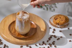 used hand pouring creamy milk into a brewing iced coffee is served with cookies. Cold coffee drink glass with ice and cream milk on white table.