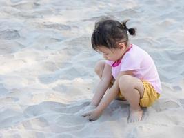 Positive charming 4 years old cute baby Asian girl, little preschooler child playing on sand beach with little wood stick on beautiful sunny day in summer time photo