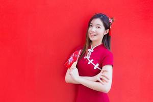 hermosa mujer asiática usa un cheongsam rojo y sostiene un ventilador rojo mientras mira a la cámara y sonríe alegremente con el fondo rojo. foto