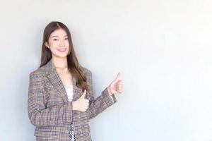 Asian professional woman with black long hair wearing a plaid suit and pretty smiling looking at camera while present product thumbs up means good on white background.