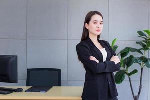 hermosa mujer de negocios asiática con un traje negro éxito de pie con los brazos cruzados en la oficina de trabajo felizmente. foto