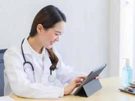 Professional beautiful Asian woman doctor searching information about patient on her tablet with happily face in the office at hospital. health care,new normal and coronavirus protection concept. photo