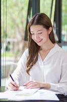 Asian confident woman wear a white shirt is using a notepad to work and there are many documents on the table. her face with smiling in a working room at home. photo