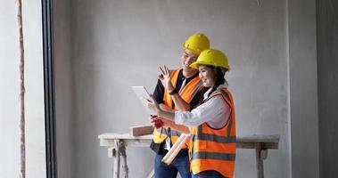 From work at a building site indoors, a young female and male foreman wearing orange vests and safety helmets are video calling on their tablets.