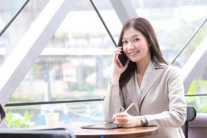Beautiful young Asian professional business female in a cream-colored suit looking at camera holds a smartphone to talk, chat and record information happily on a tablet while in a coffee shop. photo