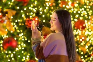 hermosa mujer asiática en un suéter colorido se encuentra feliz. en su mano sostenía una luz frente al árbol de navidad. con bokeh como fondo en el tema de las celebraciones de navidad y año nuevo foto